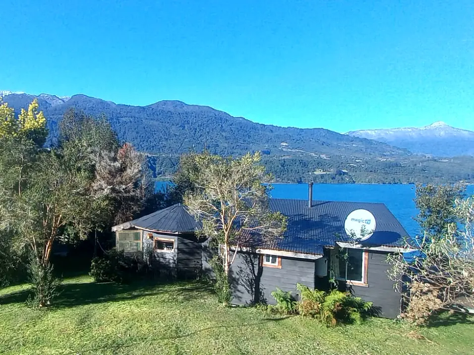 Dos cabañas de madera entre el bosque y el estuario, con montañas al fondo